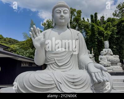 Ein beeindruckender Blick auf eine große Buddha-Statue aus Stein, die sich am historischen Ishiteji-Tempel in Matsuyama, Japan, befindet. Umgeben von üppigem Grün und blauem Himmel Stockfoto