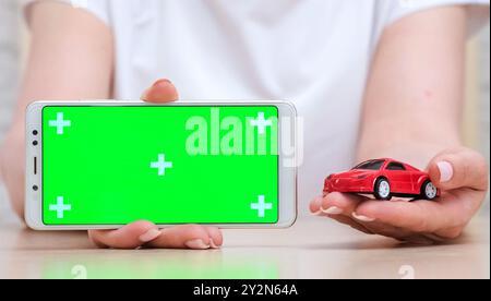 Frau mit grünem Bildschirm leeres Telefon in horizontaler Position und rotem Spielzeugauto. Anwendung zum Kaufen, Verkaufen und Mieten von Autos Stockfoto