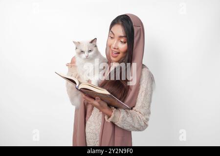 Attraktive junge asiatische muslimische Frau im Schleier-Hijab lächelt, während sie ein weißes Ragdoll-Katzentier umarmt und Al Quran auf der anderen Seite hält Stockfoto