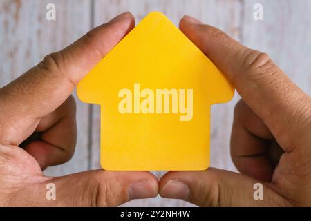 Konzeptionelles Bild mit Händen, die gelbe Hausform halten - repräsentiert Immobilienbesitz und Wohnungsmarkt. Stockfoto