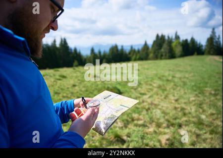 Der Mensch hält Karte und Kompass, steht auf grünem Feld mit Bäumen und Bergen im unscharfen Hintergrund. Touristen schauen sich die Karte an und navigieren durch die malerische Landschaft unter teilweise bewölktem Himmel. Stockfoto