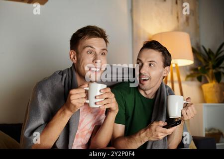 Zwei Männer teilen sich fröhliche Momente unter einer warmen Decke, während sie ihre Getränke schlürfen. Stockfoto