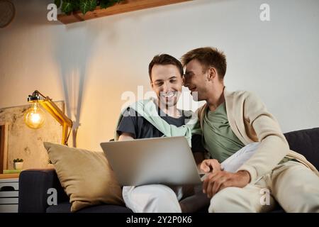 Zwei Männer teilen fröhliche Momente zusammen, während sie einen Laptop auf ihrem Sofa benutzen. Stockfoto