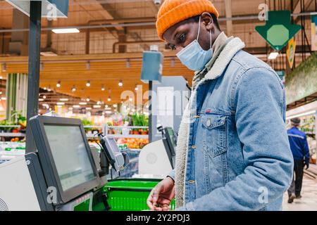 Afroamerikanischer Mann, der medizinische Maske und Denim-Jacke trägt, benutzt Selbstbedienungsmaschine im Lebensmittelgeschäft während er sicheres Einkaufen übt. Der Kunde kauft Lebensmittel und Waren im Supermarkt Stockfoto