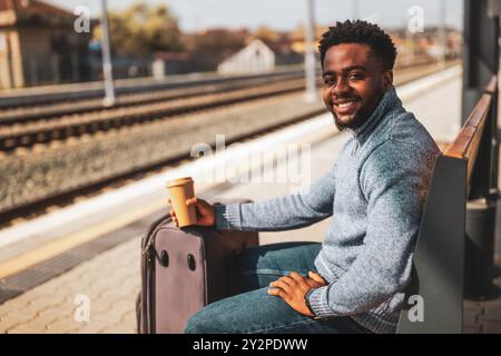 Glücklicher Mann mit Koffer trinkt Kaffee, während er auf einer Bank am Bahnhof sitzt. Stockfoto