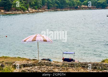 Porec, Istrien, Kroatien - 26. August 2024: Ein idyllischer Badeplatz mit Sonnenschirm und Liegestühlen an der felsigen Küste der Adria in Kroatien *** ein idyllischer Badeplatz mit Sonnenschirm und liegen an der felsigen Küste der Adria in Kroatien Stockfoto