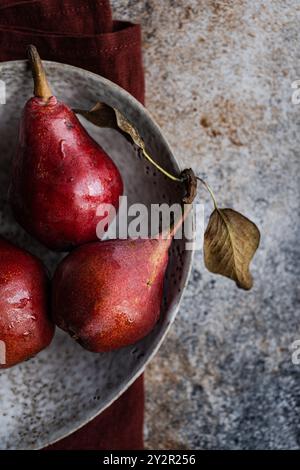 Drei frische rote Birnen aus biologischem Anbau in einer alten Keramikschale, begleitet von einem roten Tuch auf einem strukturierten Steinhintergrund Stockfoto