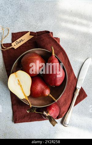 Eine Schüssel mit frischen roten Birnen, eine in zwei Hälften geschnitten, auf einer strukturierten Oberfläche mit einer rustikalen Serviette und Birnenanhänger Stockfoto
