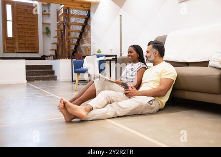 Schwarze Frau und indischer Mann sitzen auf dem Boden in einem modernen Wohnzimmer, mit ihrem Laptop und Tablet beschäftigt und genießen einen gemütlichen Tag zu Hause Stockfoto