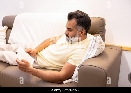 Ein indischer Mann mittleren Alters mit Bart entspannt sich in einem gemütlichen, gut beleuchteten Wohnzimmer, tief vertieft in das Lesen eines Buches Er sitzt bequem in einem Beige Stockfoto
