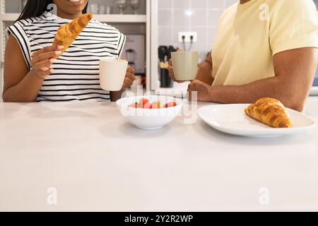 Eine schwarze Frau und ein Mann genießen ein gesundes morgendliches Essen in einer hellen Küche, bei Kaffee und Croissants mit einem Lächeln Stockfoto