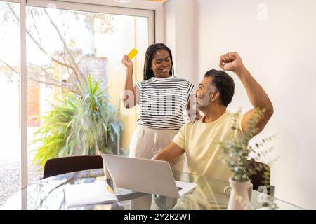Eine afroamerikanische Frau hält jubelnd eine Kreditkarte neben ihrem Partner, der triumphierend seine Faust in einem sonnendurchfluteten Home Office hochhebt Stockfoto
