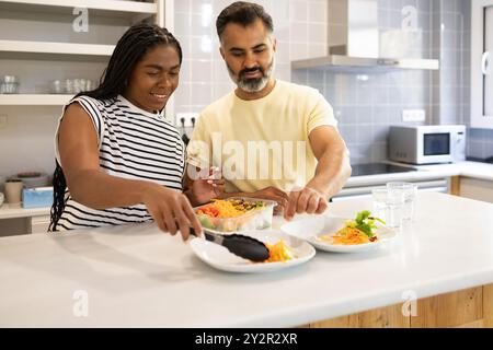 Ein Paar zwischen Rassen lächelt, während sie in ihrer modernen Küche einen frischen Salat zubereiten, der einen Moment des häuslichen Glücks und der Zusammenarbeit zeigt Stockfoto