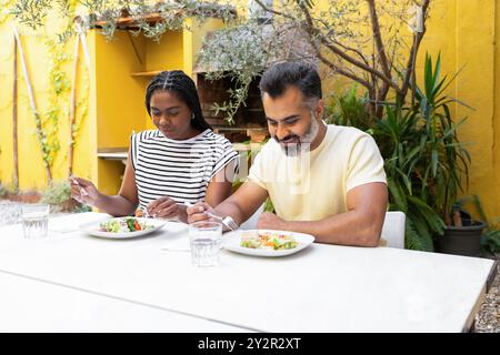 Eine schwarze Frau und ein indianer essen draußen und genießen einen sonnigen Tag in einem schönen Garten voller Pflanzen und einem warmen gelben Hintergrund Stockfoto