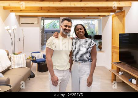 Ein fröhliches, interrassisches Paar lächelt in seinem stilvollen und geräumigen Wohnzimmer mit moderner Einrichtung und heller natürlicher Beleuchtung Stockfoto