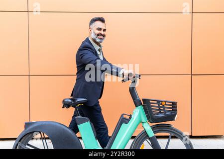 Ein Indianer im Anzug fährt mit einem Elektrofahrrad vor orangefarbenem Hintergrund durch die Stadt und bietet eine Mischung aus modernem Transport und Beruf Stockfoto