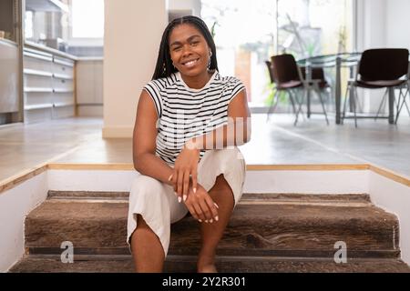 Eine fröhliche afroamerikanische Frau sitzt bequem auf Holztreppen in einem stilvollen, gut beleuchteten Haus, trägt ein gestreiftes Oberteil und eine weiße Hose und porträtiert Stockfoto