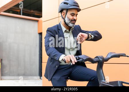 Ein Indianer in Geschäftskleidung und Helm überprüft seine Uhr, während er auf einem Elektrofahrrad in einer Stadt sitzt. Stockfoto