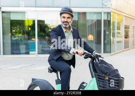 Ein indischer Mann in formeller Geschäftskleidung lächelt, während er mit einem Elektrofahrrad in einer städtischen Umgebung fährt und wegblickt. Stockfoto