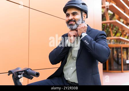 Ein Indianer, der in einem formellen Anzug gekleidet ist, passt seinen Helm im Freien an, während er neben einem Elektrofahrrad in einer städtischen Umgebung sitzt. Stockfoto