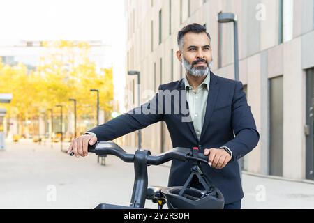 Ein professionell gekleideter Indianer steht selbstbewusst mit einem Elektrofahrrad und blickt von der Kamera weg in einer modernen Stadtlandschaft. Stockfoto