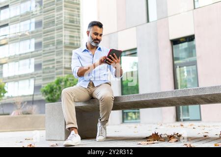 Indischer Geschäftsmann konzentrierte sich auf Tablets, während er auf einer Bank in einer modernen Stadtumgebung saß und in Business-Freizeitkleidung gekleidet war. Stockfoto