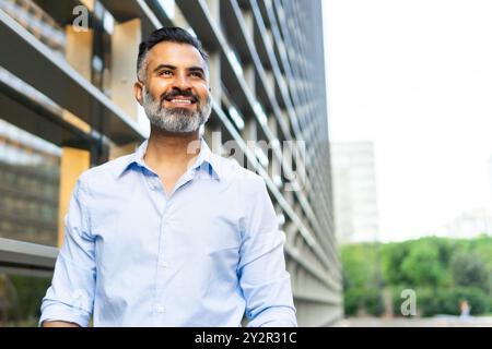 Indischer Geschäftsmann in einem formellen blauen Hemd, lächelnd und wegschauend, im Freien stehend mit moderner Gebäudekulisse in der Stadt. Stockfoto