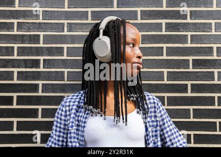 Eine schwarze Frau, die Kopfhörer trägt, genießt Musik vor einer Backsteinmauer im Geschäftsviertel und zeigt urbanen Stil und Entspannung Stockfoto
