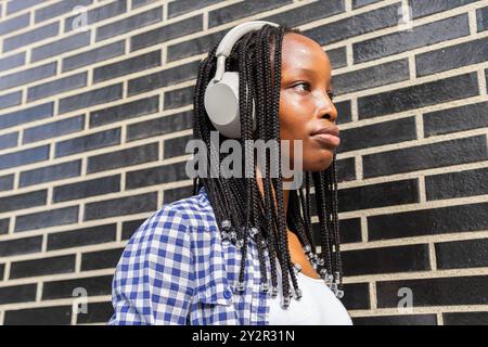 Eine junge schwarze Frau genießt Musik mit weißen Kopfhörern in einer städtischen Umgebung, lehnt sich an eine schwarze Ziegelwand. Sie wirkt besinnlich und entspannt. Stockfoto