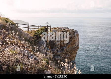 Malerischer Blick auf eine zerklüftete Klippe mit einem Holzzaun mit Blick auf das Meer in Asturien, Spanien, hervorgehoben durch sanftes Sonnenlicht und blühende Wildblumen Stockfoto