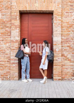 Zwei fröhliche multiethnische Studenten unterhalten sich neben einer hohen Holztür, jede trägt Taschen, die eine lässige Back-to-College-Szene widerspiegeln Stockfoto