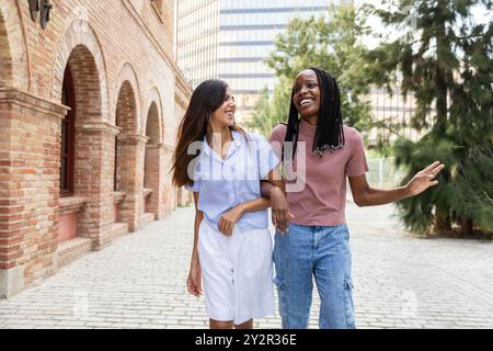 Zwei multiethnische Studenten, die einen Spaziergang auf ihrem Universitätscampus machen, plaudern und lachen, mit historischen Gebäuden im Hintergrund Stockfoto