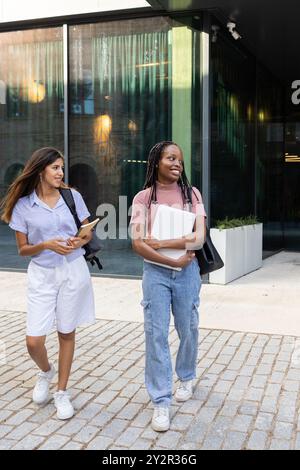 Zwei junge Studentinnen gehen zusammen auf einem Universitätscampus und unterhalten sich mit Büchern und einem Laptop in der Hand, in lässiger Atmosphäre gekleidet Stockfoto