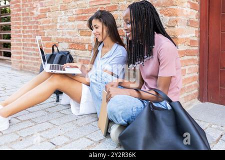Zwei Studenten, einer mit Laptop, studieren zusammen an einer Ziegelwand und verkörpern eine ungezwungene und fokussierte Bildungsumgebung Stockfoto