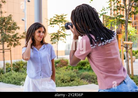 Eine junge Studentin posiert für eine Fotosession auf einem sonnendurchfluteten College-Campus, die von einer anderen Studentin mit einer Kamera aufgenommen wird und Kreativität und Kreativität zum Ausdruck bringt Stockfoto