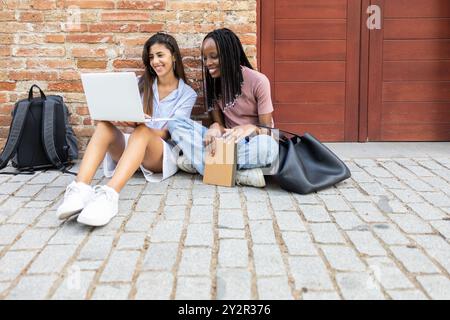 Zwei verschiedene Studentinnen sitzen draußen auf einem Kopfsteinpflasterweg vor einer Mauer, lernen zusammen mit einem Laptop, lächeln und beteiligen sich Stockfoto