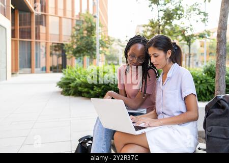 Zwei junge Frauen studieren draußen auf einem Universitätscampus und benutzen einen Laptop auf einer Bank, die von modernen akademischen Gebäuden umgeben ist Stockfoto