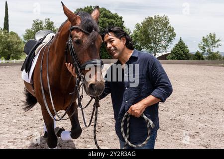 Ein Bräutigam pflegt sanft ein braunes Pferd mit Reitausrüstung in einer Sandarena. Die Umgebung spiegelt eine ruhige Verbindung zwischen Mensch und Tier inmitten von lu wider Stockfoto