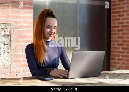 Eine junge Frau mit rotem Haar, mit blauem Rollkragen, sitzt draußen an einem Holztisch, auf ihren Laptop gerichtet, mit einem Smartphone neben ihr. Stockfoto