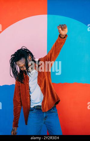 Ein fröhlicher afroamerikanischer Mann tanzt energisch vor einem hellen, bunten Hintergrund, seine langen Dreadlocks und die leuchtende orange Jacke ergänzen ihn Stockfoto