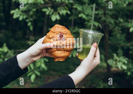 Ein nicht wiedererkennbares Teenager-Mädchen, das ein leckeres Croissant in Kombination mit einem erfrischenden Sommer-Matcha-Getränk genießt, in einer natürlichen Umgebung im Freien. Stockfoto