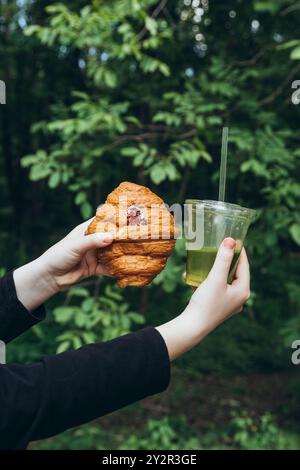Ein nicht wiedererkennbares Teenager-Mädchen, das ein Croissant und ein eiskaltes Matcha-Getränk in üppigem Grün hält. Das Foto konzentriert sich auf das Essen und Trinken, die Herstellung Stockfoto