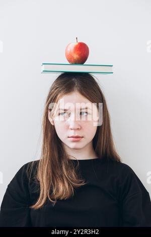 Ein Mädchen im Teenageralter, das mit einem neutralen Ausdruck von der Kamera wegschaut und ein Buch und einen hellroten Apfel auf dem Kopf vor einem schlichten Hintergrund ausbalanciert. Stockfoto