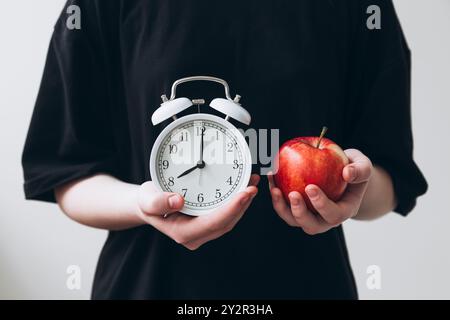 Beschnittene, nicht erkennbare Person hält einen weißen Wecker und einen roten Apfel. Stockfoto