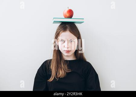 Ein Teenager-Mädchen, das einen Apfel auf Büchern auf dem Kopf balanciert, steht vor einem einfachen Hintergrund und starrt direkt in die Kamera. Stockfoto