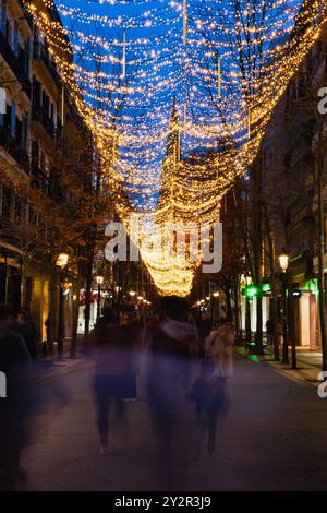 Eine Stadtstraße, die mit faszinierenden blauen und gelben Weihnachtslichtern beleuchtet ist und am Abend ein festliches Baldachin über den unscharfen Fußgängern schafft Stockfoto