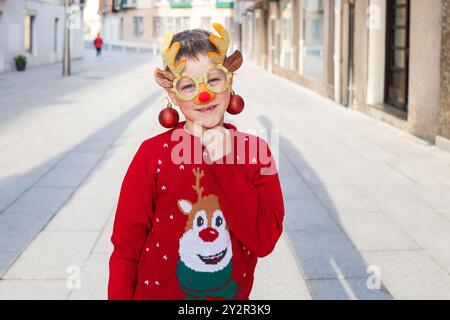 Ein fröhlicher kleiner Junge mit Rentierbrille und rotem Weihnachtspullover steht draußen und verkörpert den festlichen Geist der Weihnachten Stockfoto