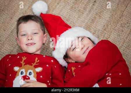 Ein kleiner Junge und ein Mädchen, beide mit festlichen Weihnachtsmützen und gemütlichen Weihnachtspullover, liegen Seite an Seite auf einem Teppich, lächeln fröhlich und verkörpern die Wa Stockfoto