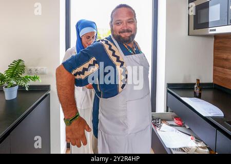 Zwei Freunde teilen sich einen freudigen Moment in der Küche, einer in einer traditionellen Schürze, der andere in Kulturkleidung und bereiten sich lachend auf ein Barbecue vor Stockfoto