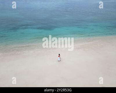 Aus der Vogelperspektive einer Frau in einem fließenden weißen Kleid, die allein an der unberührten Küste eines abgelegenen Strandes in Mauritius spaziert, umgeben von pulsierendem Türkis Stockfoto